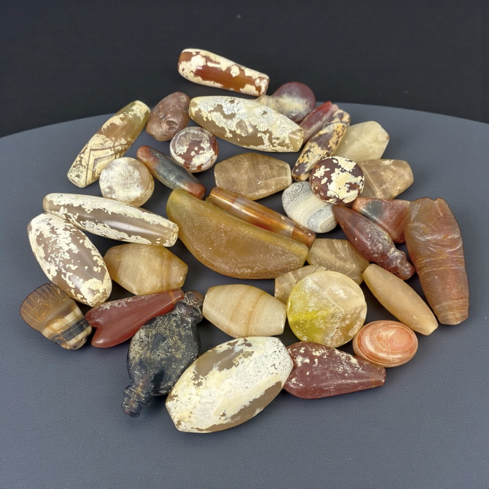 A Group of Parthian-Sasanian Agate Beads, Circa 1st-7th Century CE, 9W: The group comprises thirty-four agate beads of varying forms and dimensions, carved from banded and translucent chalcedony in tones of brown, amber, cream, red, and grey. The beads include elongated t