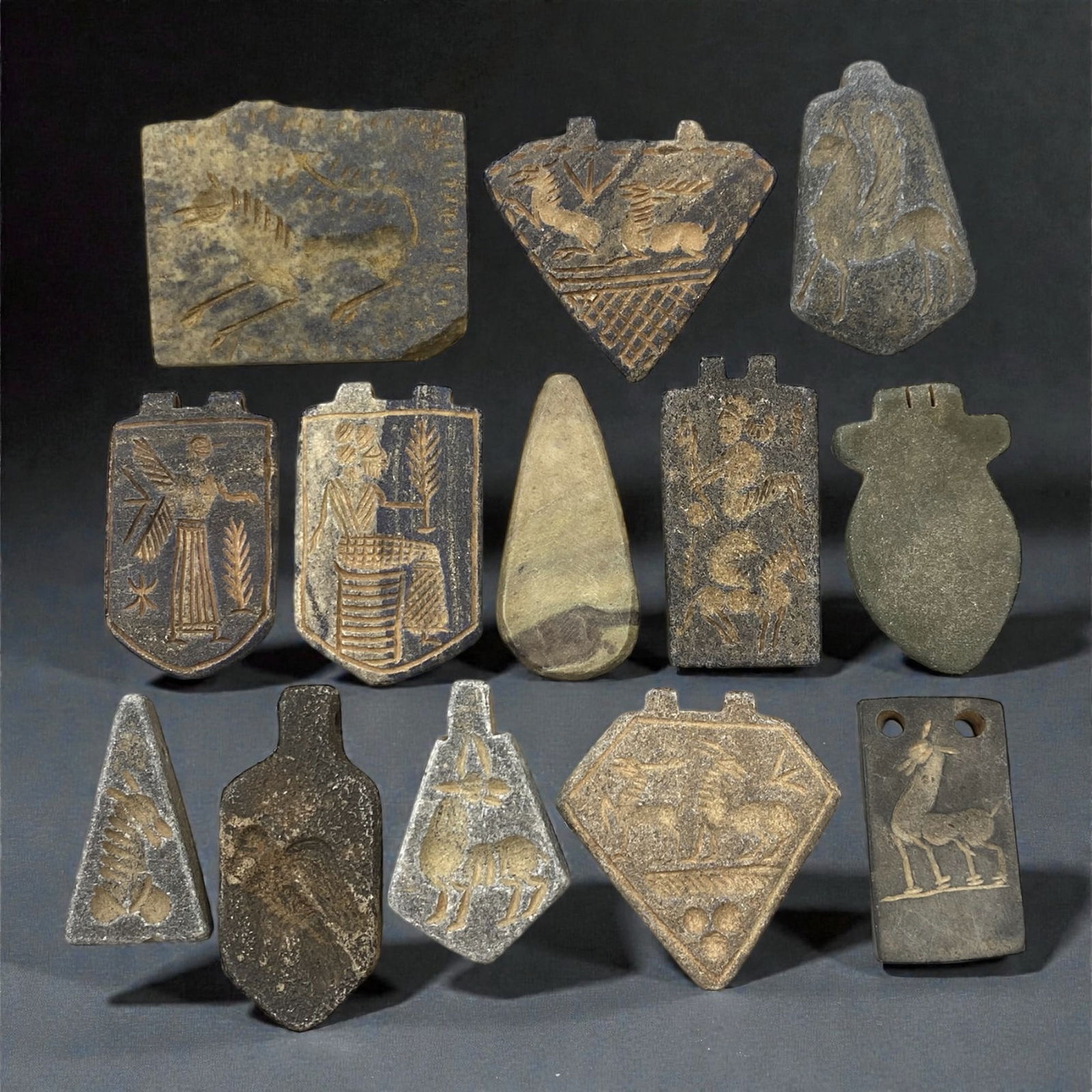 A Group of Near Eastern Stone Amulets, 1st Millennium BCE, 70W: The group comprises thirteen stone amulets carved from grey, buff, and dark-toned stone, including steatite and related materials. The amulets range in form and include rectangular, trapezoidal, trian