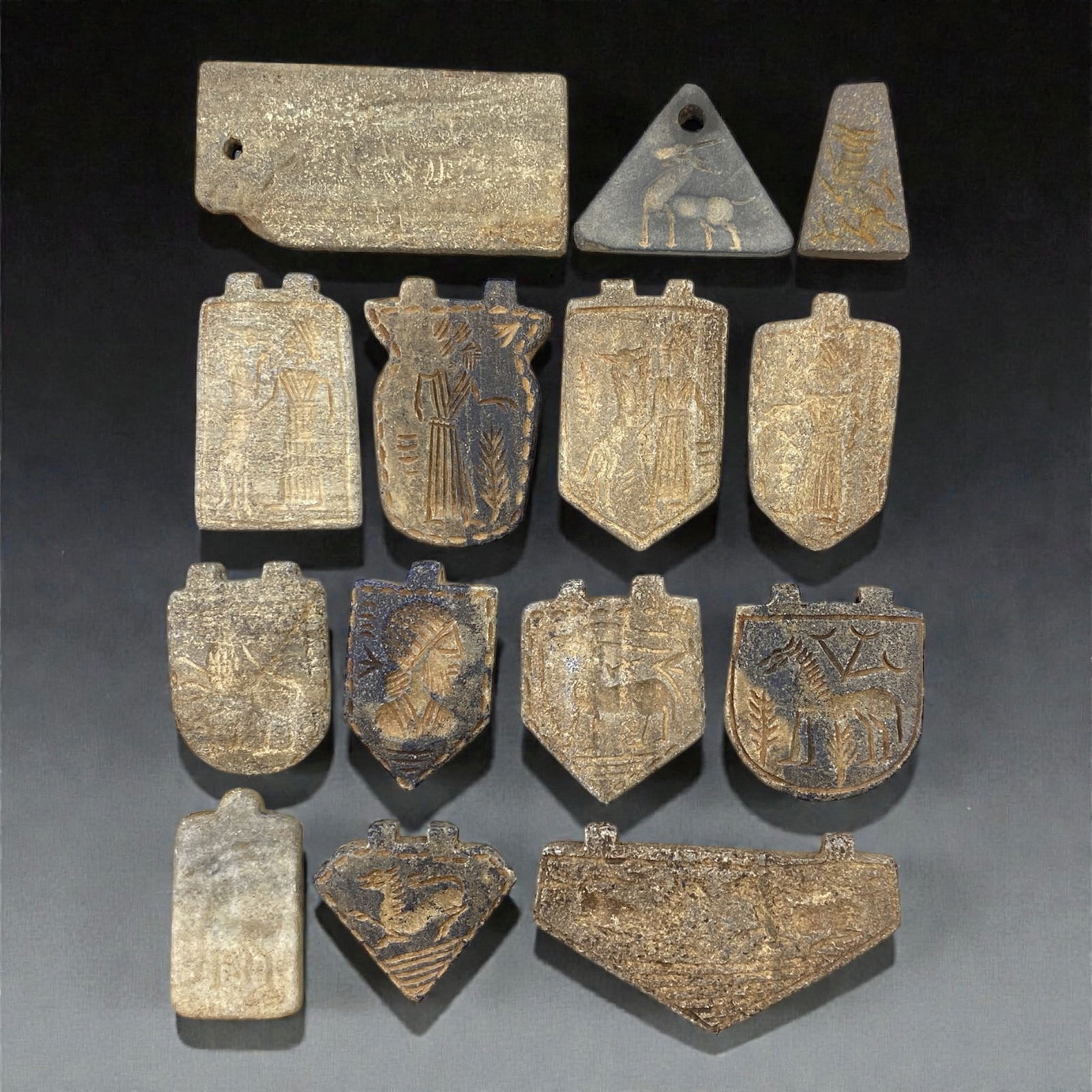 A Group of Near Eastern Stone Amulets, 1st Millennium BCE, 60W: The group comprises fourteen stone amulets carved from grey, buff, and dark-toned stone, including steatite and related materials. The amulets vary in size and shape and include rectangular, shield-sh