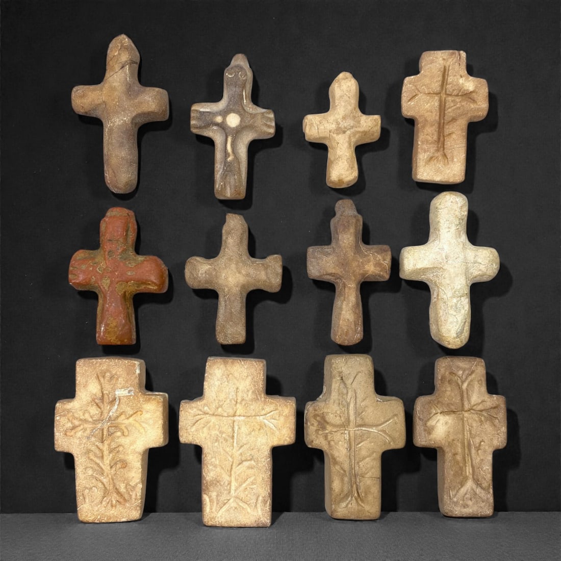 A Group of Medieval Christian Stone Crosses, 12th-15th Century, 66W: Carved from light beige, buff, reddish, and grey stone with natural inclusions, veining, and areas of surface wear, the group comprises twelve small stone crosses executed in simple relief. The crosse