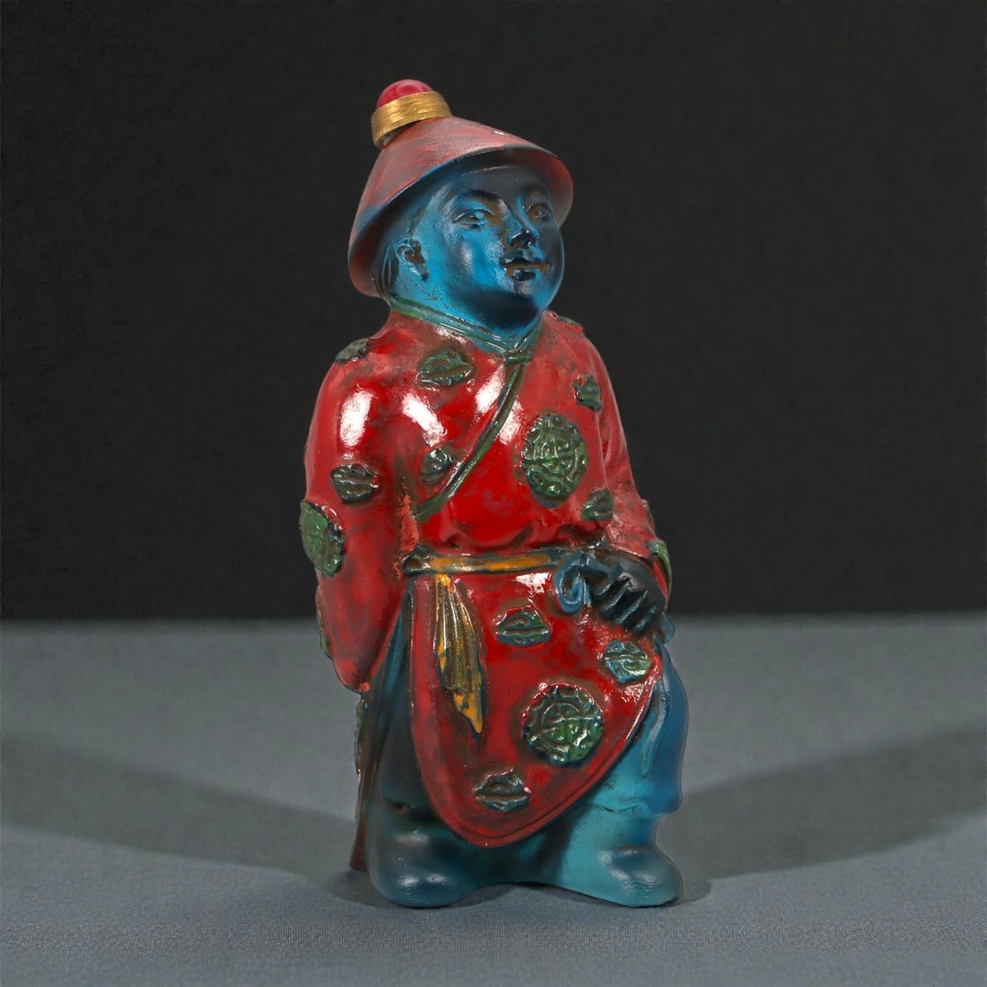 A Chinese Painted Glass Figural Snuff Bottle, Qing Dynasty, Late 19th Century, 6YB: Molded from translucent blue glass, the bottle is formed in the round as a standing male figure wearing a long robe and a brimmed hat. The facial features are softly modeled, with rounded cheeks, almo