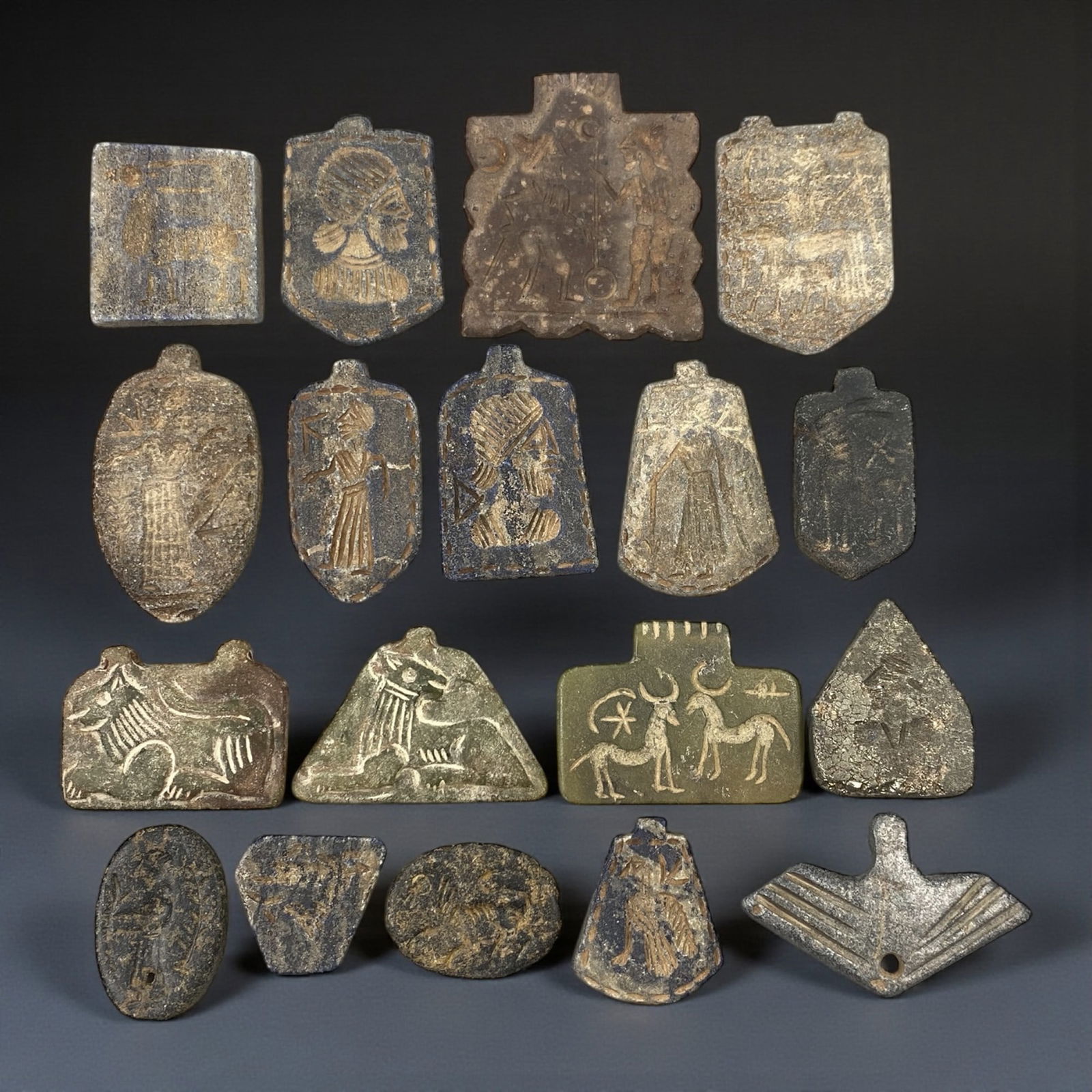 A Group of Near Eastern Stone Amulets, 1st Millennium BCE, 61W: The group comprises eighteen small stone amulets carved from grey, buff, and dark-toned stone, including steatite and comparable soft stones. The objects display a range of forms, including rectangula