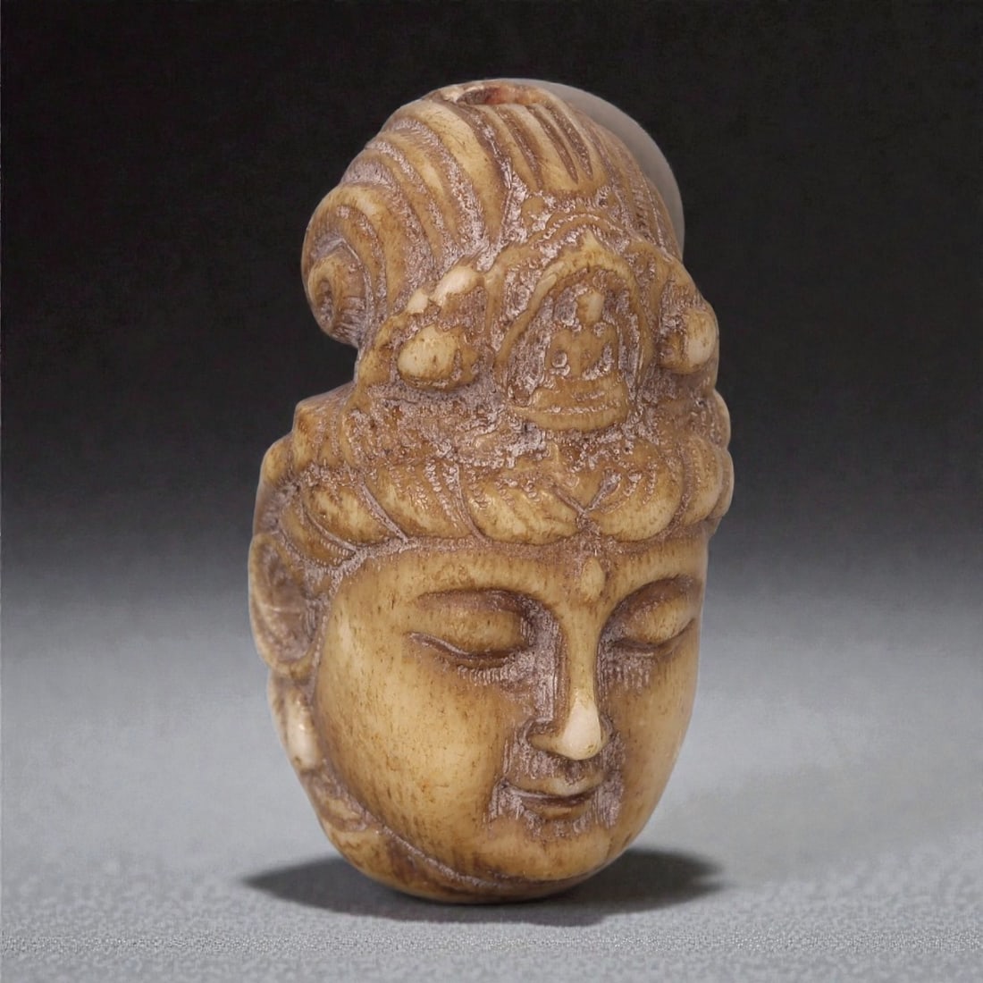 A Chinese Bone Bead Carved as a Buddhist Head, 19th-20th Century, 100FC: Carved from bone with a warm beige tone and areas of darker patination, the bead is modeled in the form of a Buddhist head shown frontally. The face is rendered with closed eyes, gently arched brows,
