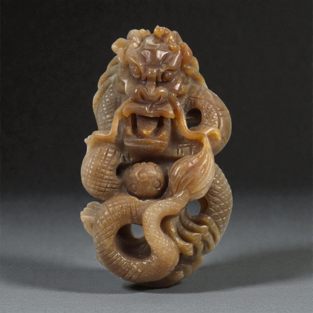 A Chinese Nephrite Jade Dragon Pendant, 20th Century, 33FB: Carved from warm brown nephrite jade with areas of lighter translucency, the pendant is worked in openwork relief as a coiled dragon shown grasping a pierced sphere. The dragon is rendered with a styl