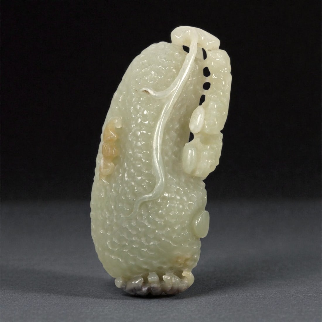 A Chinese Nephrite Jade Fruit-Form Pendant, 20th Century, 65FA: Carved from pale celadon nephrite jade with areas of translucency and minor russet inclusions, the pendant is modeled in the form of an elongated fruit. The surface is densely worked with small rounde