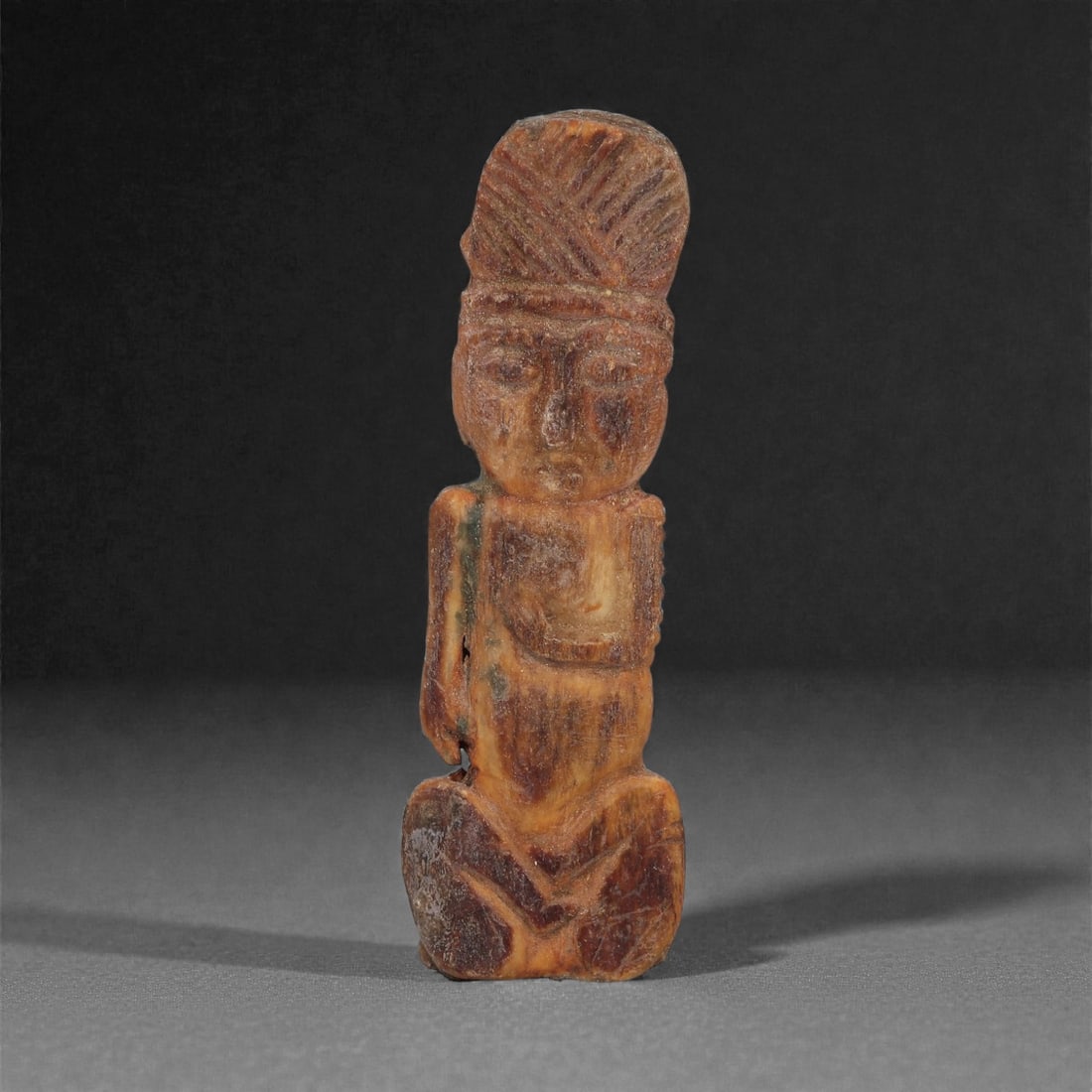 A Coptic Carved Bone Figure, 5th-7th Century CE, 11LJ: Carved from a warm brown bone with darker surface accretions, the figure is shown seated with the legs folded beneath and the arms held close to the body. The head features a tall, striated headdress,