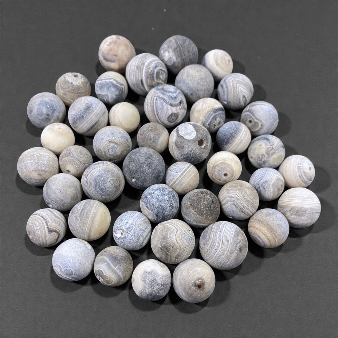 A Group of Tibetan Agate Beads, 18th-19th Century, 60V: Comprising forty-three spherical and near-spherical beads carved from agate, the group displays a restrained palette of grey, pale blue-grey, cream, and off-white tones, with many examples exhibiting