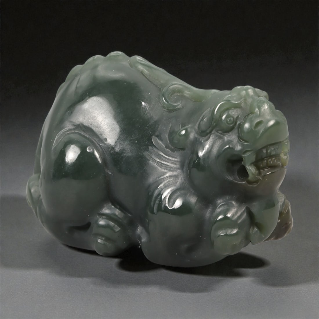 A Chinese Nephrite Jade Mythical Beast Figure, 20th Century, 64YA: Carved from dark green nephrite jade with areas of deeper saturation and soft translucency, the figure represents a recumbent mythical beast shown in a compact, crouching posture. The head is rendered