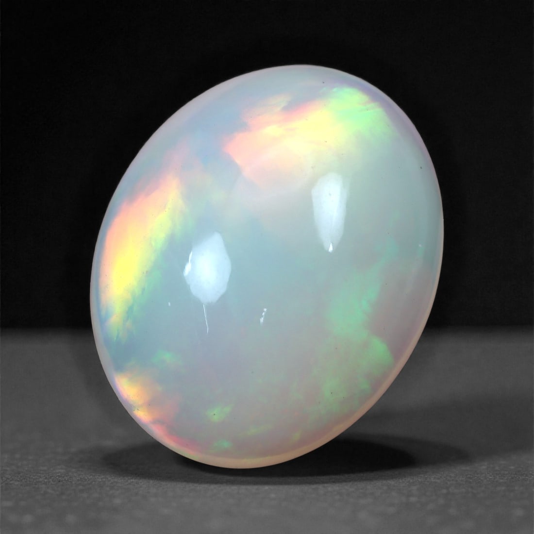 6.36 Carats Oval Cabochon Opal - Natural Ethiopian Stone 16.8x13.5x6.5mm, 6XH: This mesmerizing oval cabochon opal radiates with vibrant play-of-color, displaying bursts of lime green, neon yellow, and fiery orange that shimmer across its translucent white body. The gem's rounde