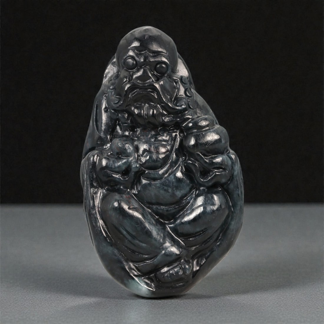 A Chinese Dark Nephrite Jade Figure of Budai, 20th Century, 39FB: Carved from dark green to near-black nephrite jade with subtle tonal variation and a high polish, the figure represents Budai shown seated with legs crossed and body drawn into a compact, rounded form