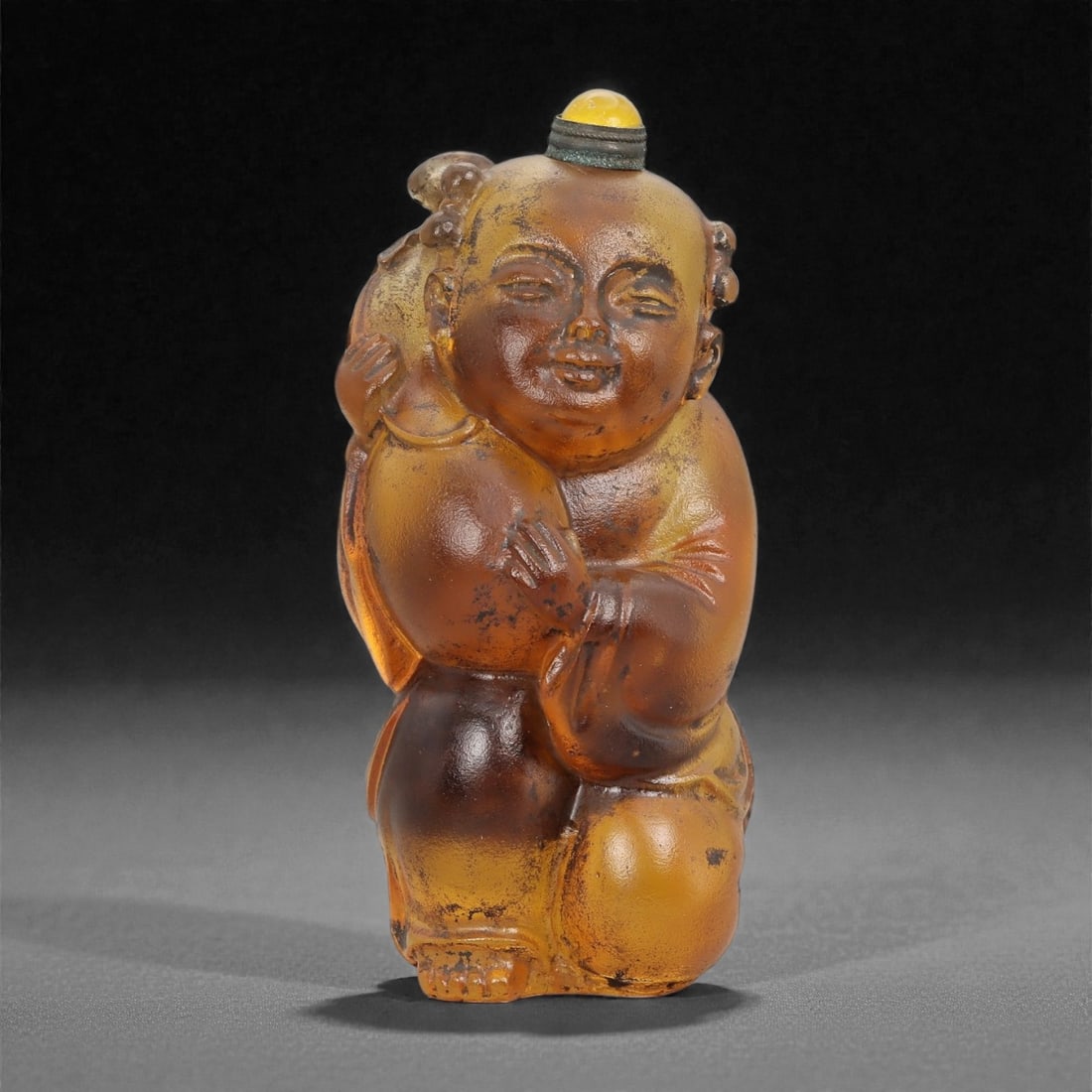 A Chinese Glass Figural Snuff Bottle, 20th Century, 70FG: Molded from translucent amber-colored glass with uneven tonal saturation and scattered dark inclusions, the snuff bottle is formed as a standing infant figure rendered fully in the round. The child is