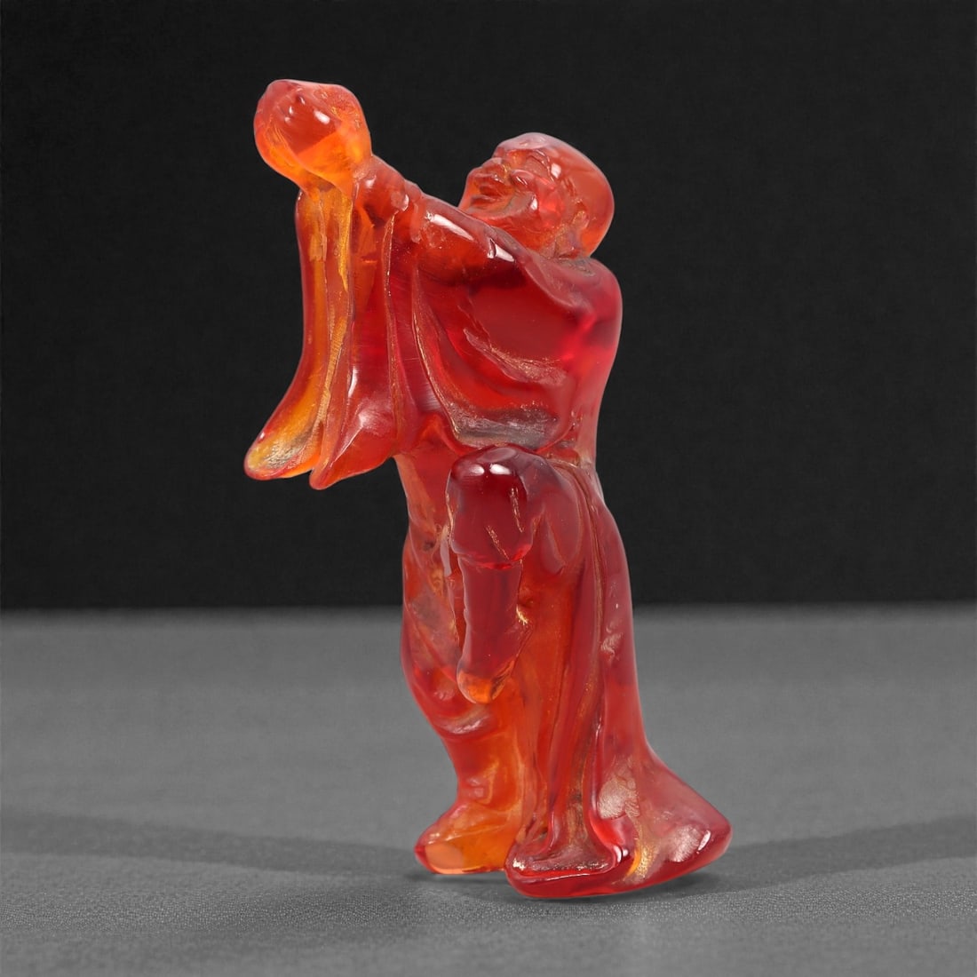 A Chinese Amber-Resin Figure of a Luohan, 20th Century, 36FH: Cast or carved from translucent amber-colored red resin with internal tonal variation, localized inclusions, and minor entrapped bubbles, the figure represents a standing luohan shown in an animated,