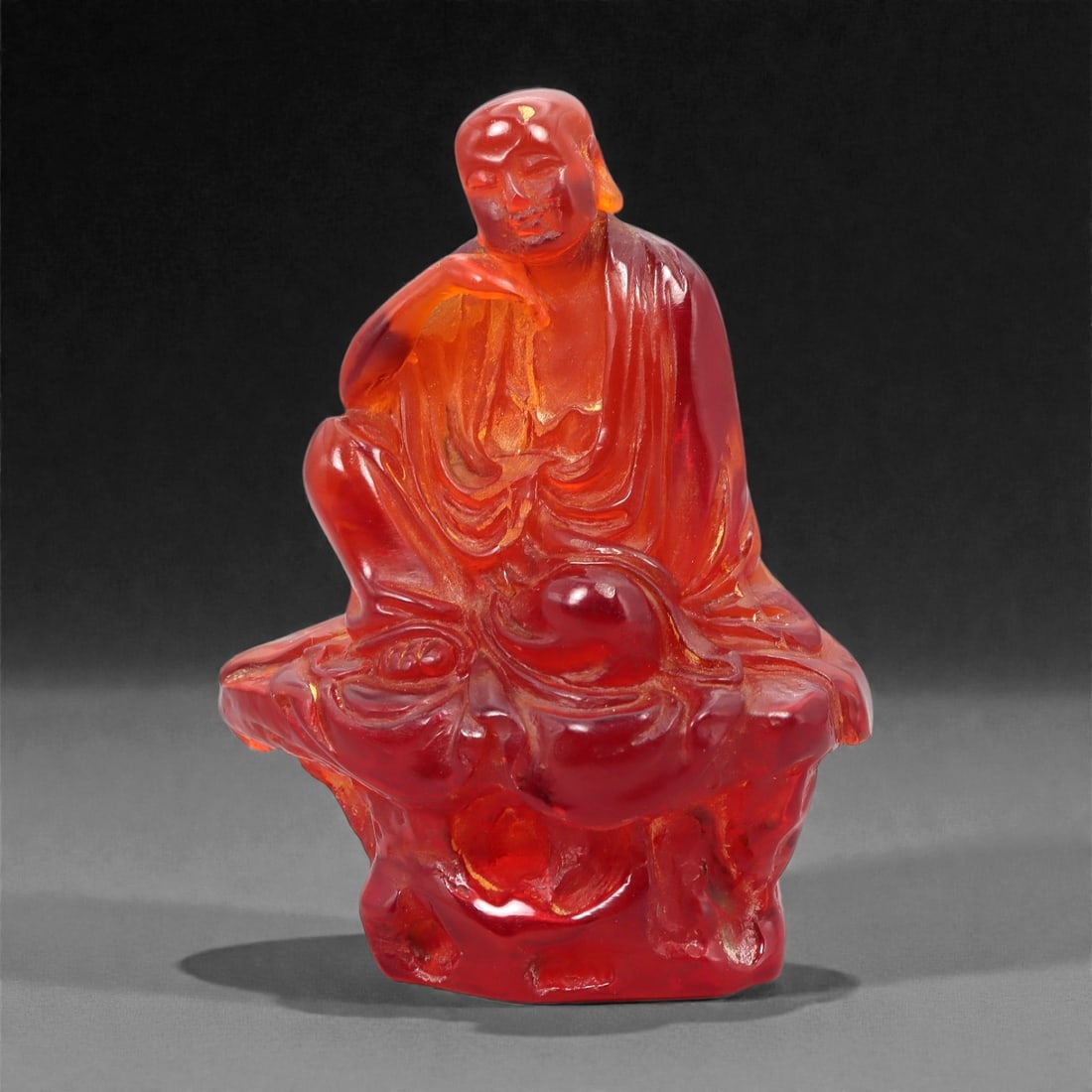 A Chinese Amber-Resin Figure of a Luohan, 20th Century, 39FH: Cast or carved from translucent amber-colored red resin with internal tonal variation, localized inclusions, and minor entrapped bubbles, the figure represents a seated luohan shown resting on a styli