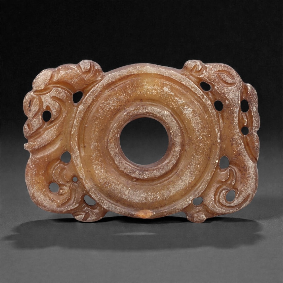 A Chinese Hetian Jade Openwork Plaque, 20th Century, 82FH: Carved from warm brown-toned Hetian nephrite jade with lighter translucent areas and natural surface mottling, the plaque is worked in an openwork format centered on a circular aperture. The surroundi