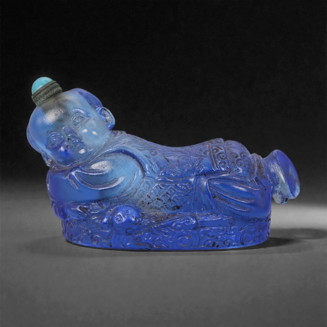 A Chinese Glass Snuff Bottle, 20th Century, 51FG: Molded from translucent cobalt-blue glass, the snuff bottle is formed as a reclining child shown resting on a low, oval base. The figure is rendered in low to moderate relief, with rounded facial feat