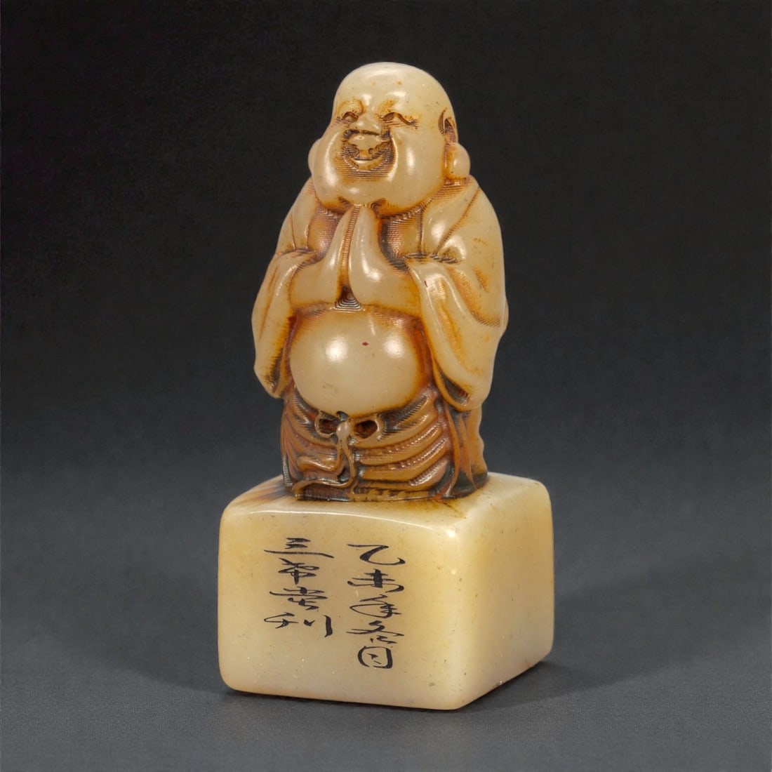 A Chinese Nephrite Jade Laughing Buddha Stamp Seal, 20th Century, 18FA: Carved from pale celadon nephrite jade with warm russet inclusions and a smooth, glossy polish, the seal is surmounted by a standing figure of the Laughing Buddha (Budai). The figure is rendered with