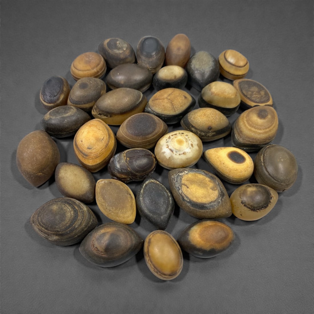 A Group of Tibetan Luk Mik ("Goat's Eye") Agate Beads, 11th-14th Century CE, 27W: Comprising thirty-three agate beads of ovoid to lenticular form, carved from naturally banded agate in tones of brown, amber, honey, grey, and black, the group is characterized by prominent concentric