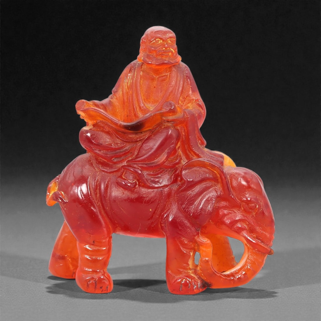 A Chinese Amber-Resin Figure of a Luohan Riding an Elephant, 20th Century, 37FH: Cast or carved from translucent amber-colored red resin with internal tonal variation and minor inclusions, the figure depicts a luohan seated astride an elephant. The luohan is shown wearing a draped