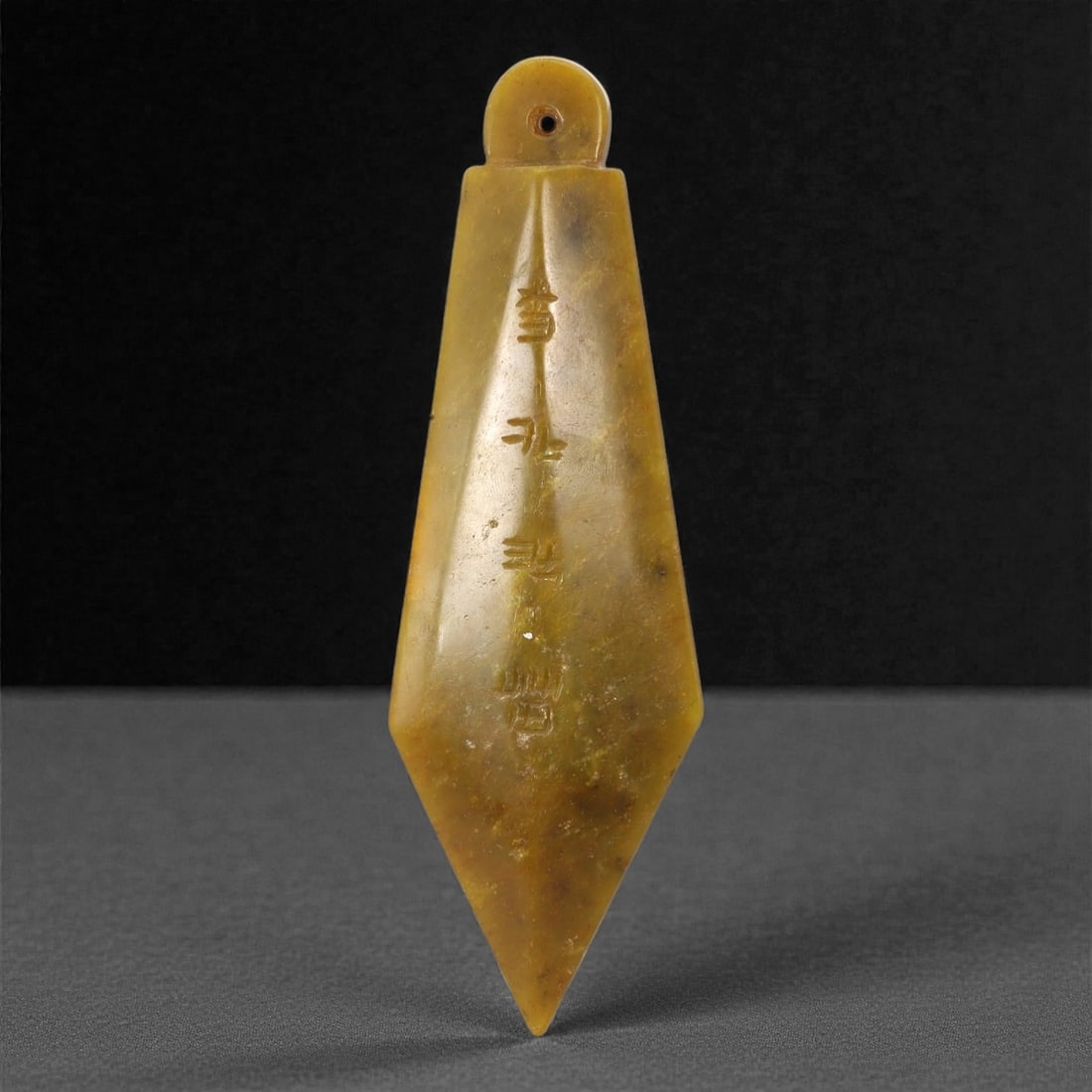 A Chinese Hetian Jade Inscribed Pendant, 20th Century, 61FH: Carved from translucent yellow-toned Hetian nephrite jade with natural clouding and darker inclusions, the pendant is worked as a tapering triangular form with a rounded upper terminal. A circular per