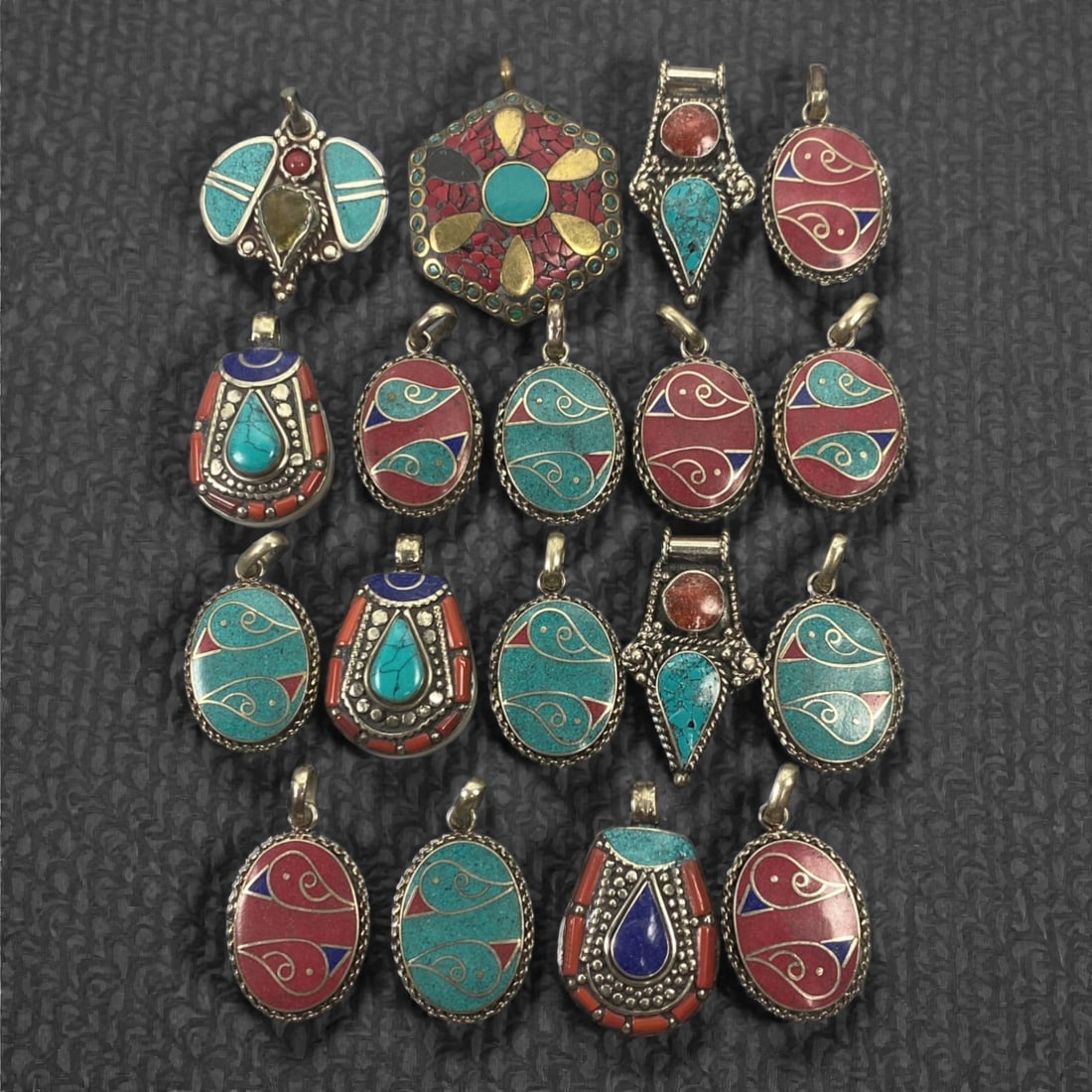A Group of Himalayan Silver-Mounted Stone Inlay Pendants, 20th Century, 98FK: The group comprises eighteen silver-mounted pendants of varying forms, including oval, teardrop, shield-shaped, and polygonal examples, each decorated with inset hardstones and composite inlay. The mo