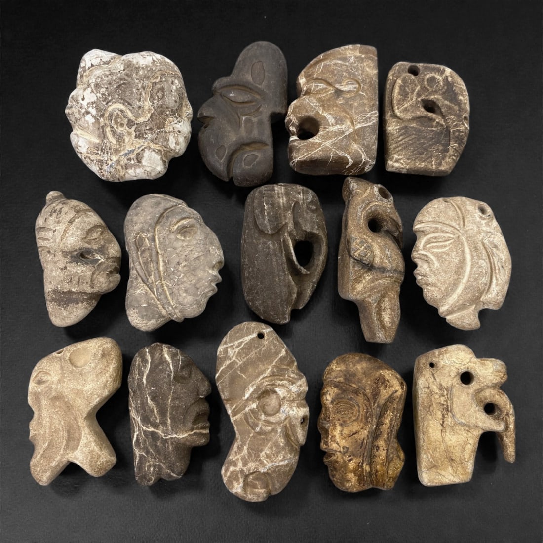A Group of Mesoamerican Stone Sculptural Pendants, Pre-Columbian Period, 77Z: Carved from various stone types in tones ranging from light grey to dark brown, the group comprises fourteen sculptural pendants representing stylized human heads and abstracted anthropomorphic forms.