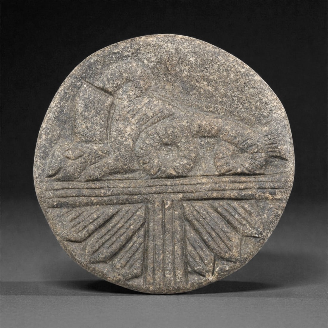 An Indo-Parthian Stone Plaque, 1st-3rd Century CE, 25ZA: Carved from a grey stone with granular surface texture, the plaque is circular in form and bears a low-relief depiction of a recumbent animal, possibly a lion, shown on a groundline articulated by hor