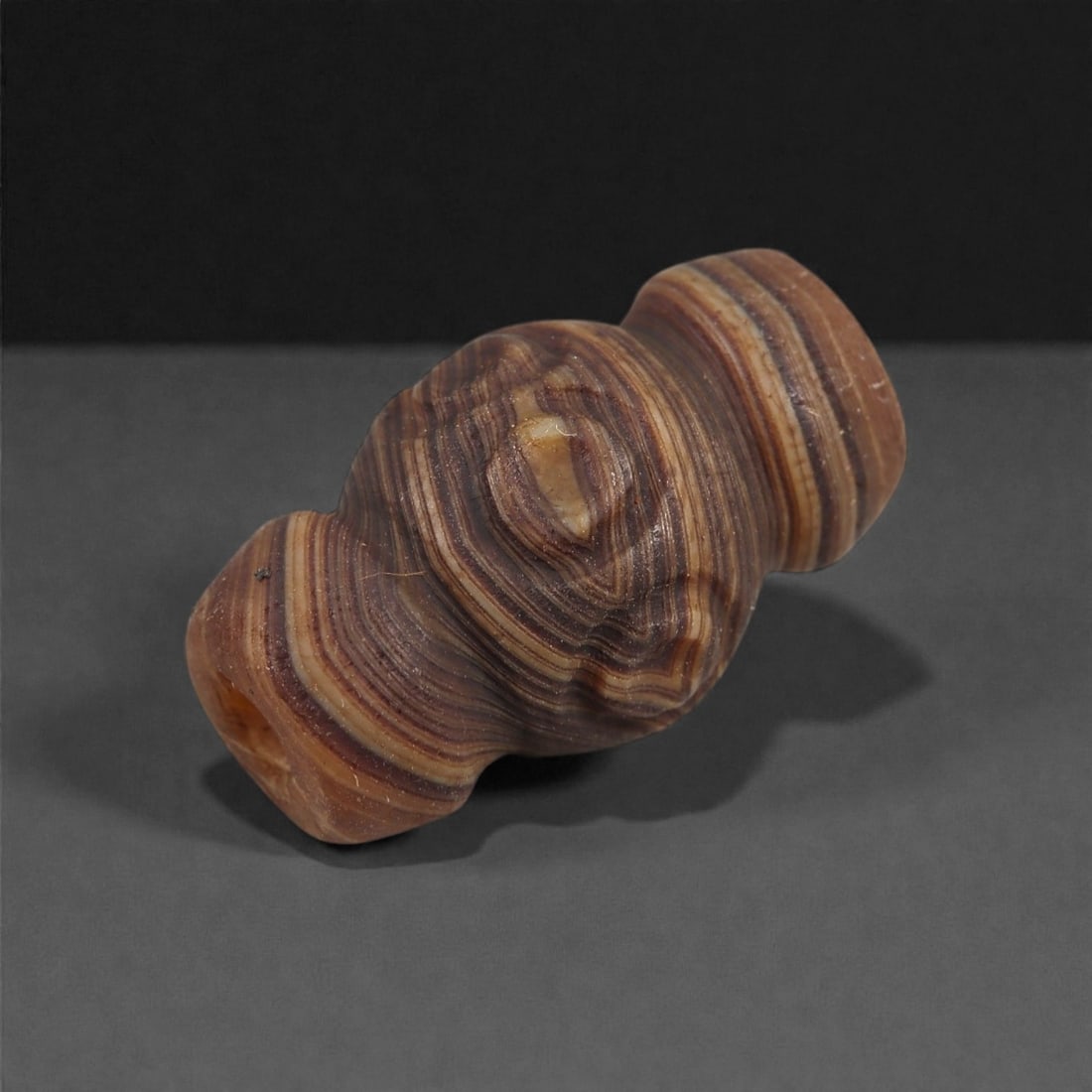 A Chinese Agate Bead, Qing Dynasty, 18th-19th Century, 71XK: Carved from banded agate displaying alternating brown, tan, and cream striations, the bead is formed as a short tubular element with gently rounded contours. The natural concentric banding of the ston