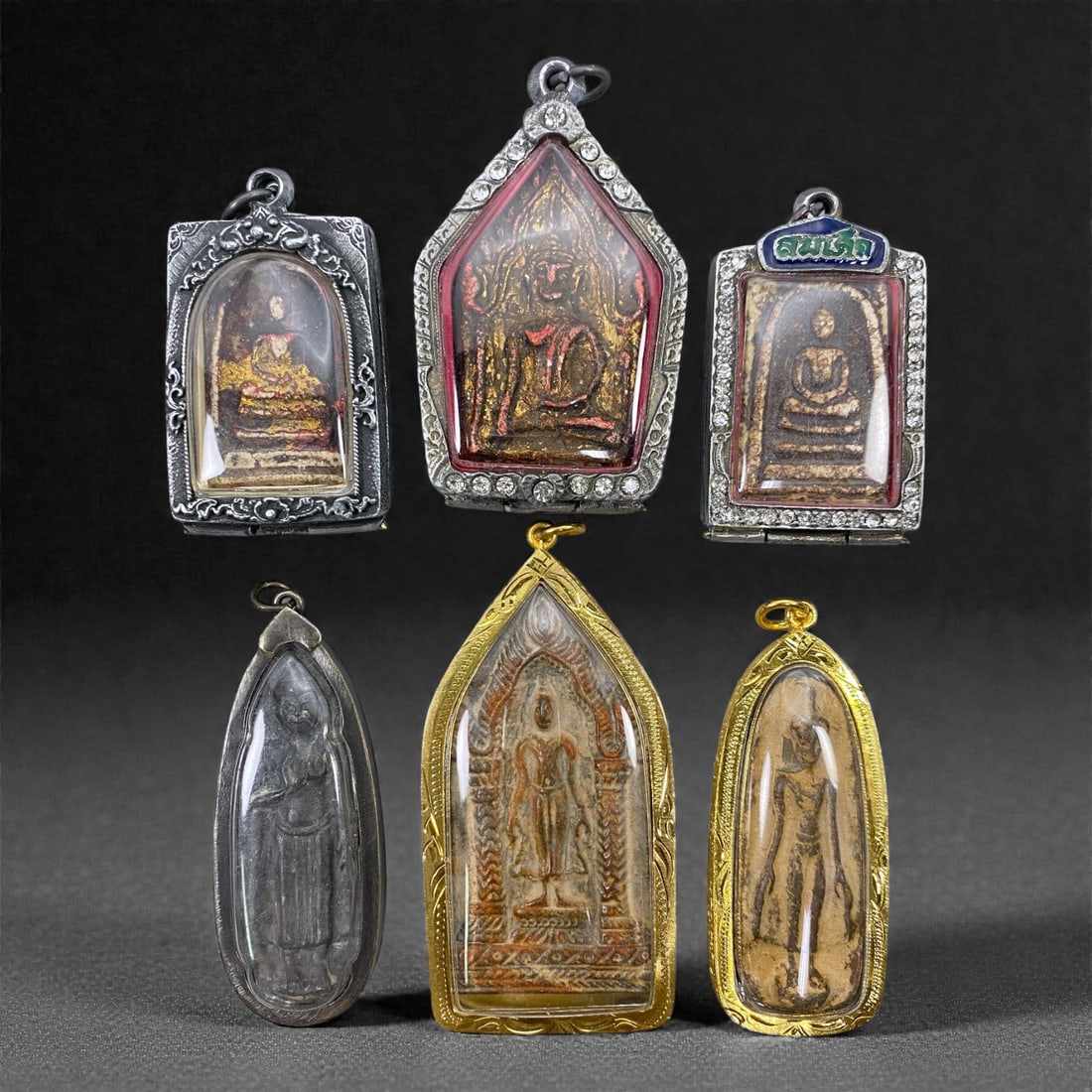 A Group of Six Thai Buddhist Votive Amulets, 20th Century, 97Z: Comprising six Thai Buddhist votive amulets of elongated and arched formats, each mounted in an individually made silver or white-metal frame with integral suspension loop, the group consists of molde
