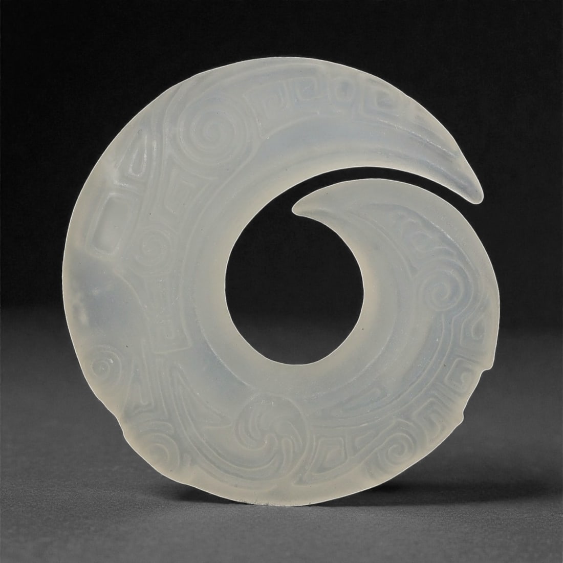 A Chinese Pale Jade Openwork Pendant, 20th Century, 92FD: Carved from translucent pale jade of even milky tone, the pendant is worked as a circular openwork form with an inward curling projection, creating a continuous flowing silhouette. The surface is deco