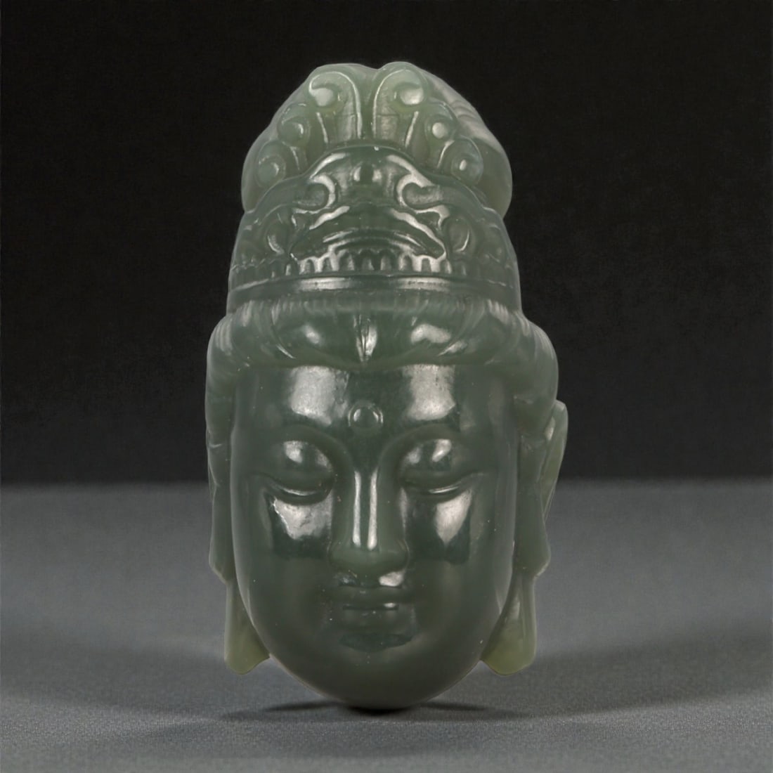 A Chinese Nephrite Jade Guanyin Head Pendant, 20th Century, 2YB: Carved from dark green nephrite jade with areas of lighter translucency and a high-polish surface, the pendant is formed as a head of Guanyin shown frontally. The face is modeled with softly rounded f