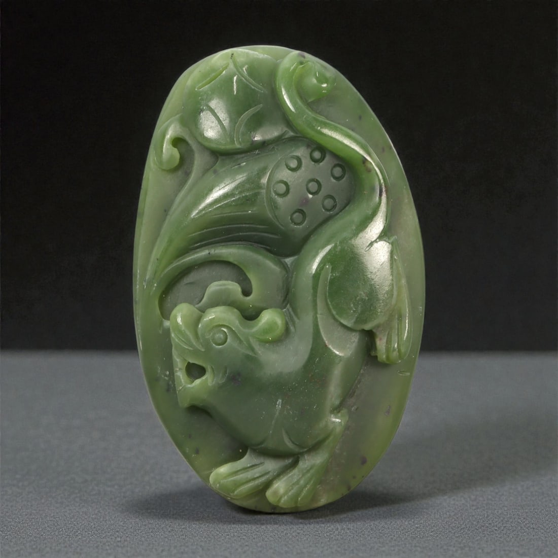 A Chinese Nephrite Jade Relief Plaque, 20th Century, 52YA: Carved from dark green nephrite jade with mottled tonal variation and scattered darker inclusions, the plaque is worked in layered relief depicting a stylized mythical beast within a compact, oval for