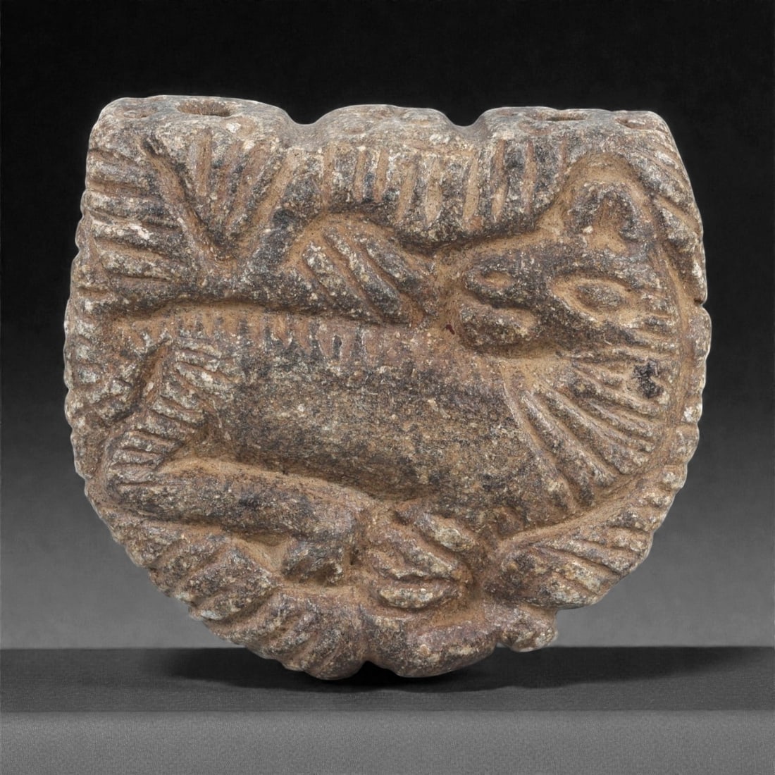 A Near Eastern Schist Zoomorphic Amulet, 2nd-1st Millennium BCE, 58LJ: Carved from dark grey schist, the amulet is worked in low relief with a reclining quadruped shown within a framing border of incised radiating strokes. The animal displays a rounded head with recessed