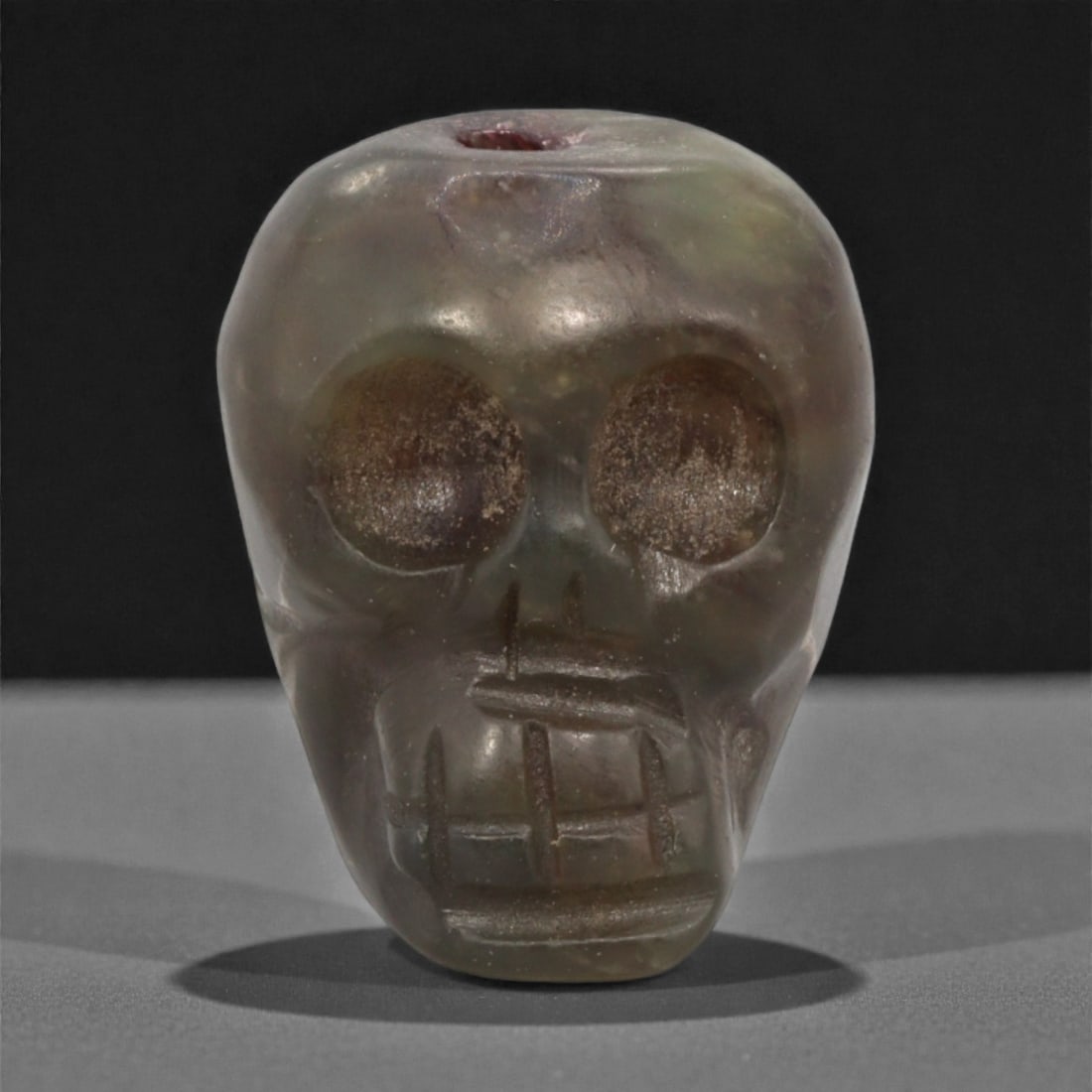 A Tibetan Stone Skeleton Bead, Mid-20th Century, 69FJ: Carved from translucent grey-green stone with darker brown inclusions and subtle internal veining, the bead is formed as a stylized human skull rendered in frontal view, with large circular recessed e