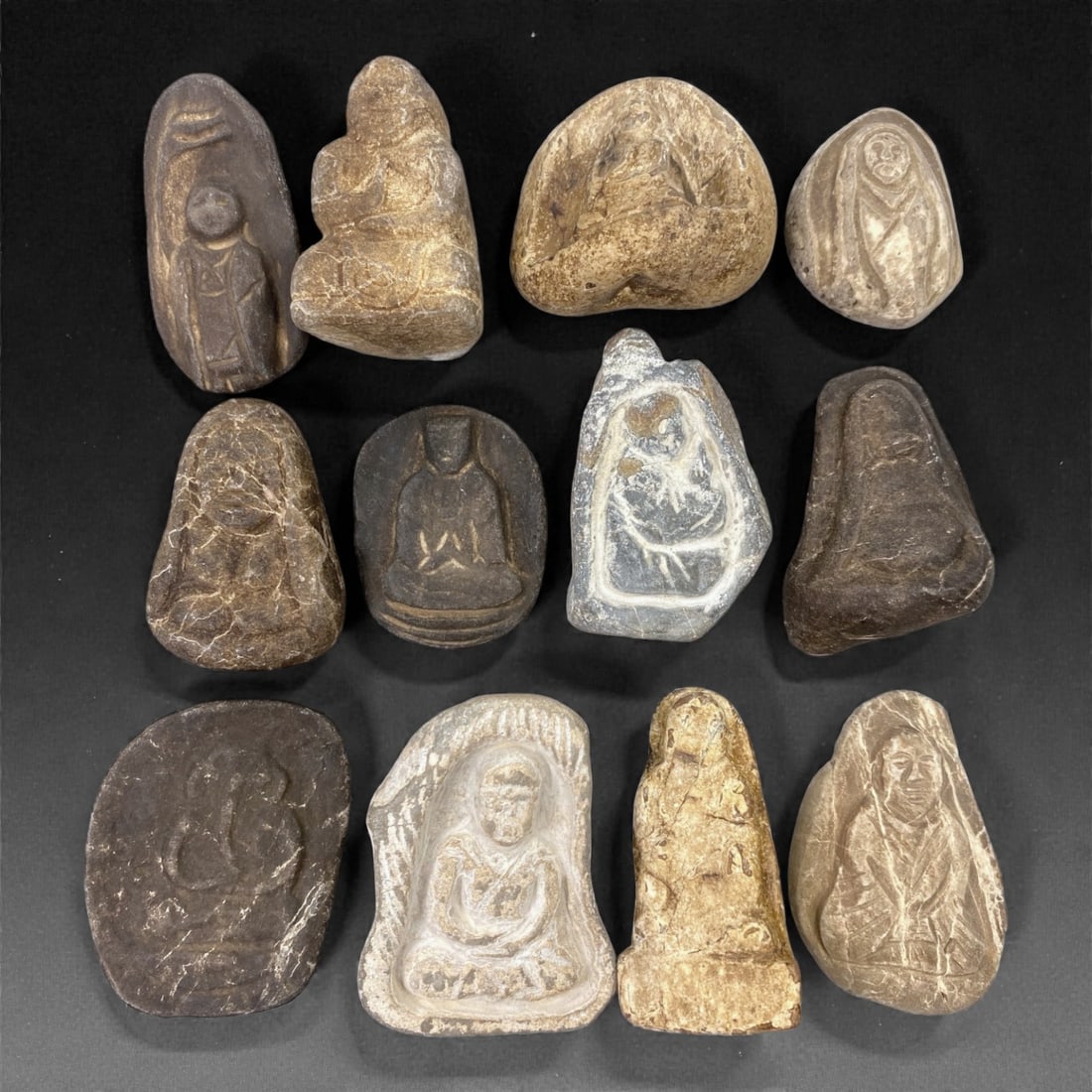 A Group of Japanese Stone Buddhist Figures, Edo Period, 92Z: Carved from light to dark-toned stone with natural veining, inclusions, and areas of surface wear, the group comprises twelve small Buddhist devotional figures of irregular, rounded, and pebble-like f