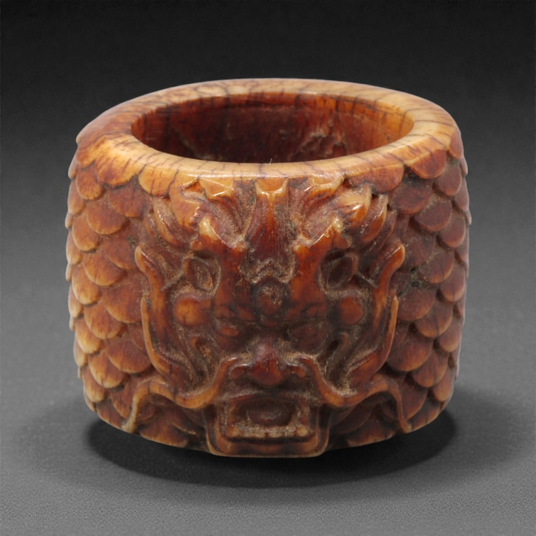 A Chinese Bone Thumb Ring with Dragon Head, 20th Century, 75FH: Carved from bone with warm brown and honey-toned coloration, the thumb ring is cylindrical in form with a thickened wall and hollow interior. The exterior is carved in high relief with a frontal drago