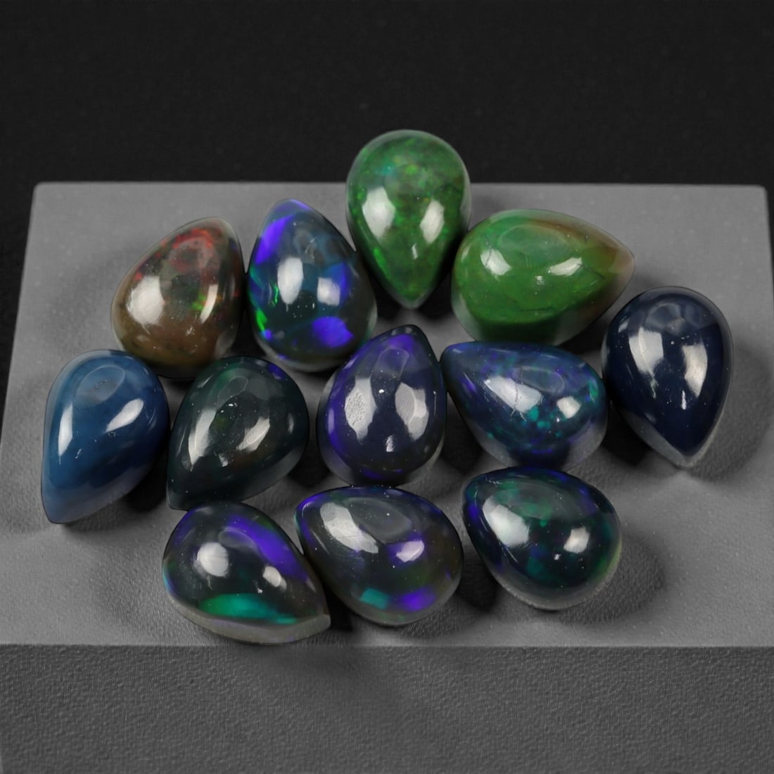 6.86 Carats Multicolor Welo Opal Teardrop Selection - Natural Ethiopian Stones, 30XK: This dazzling group of Welo opal cabochons radiates with a kaleidoscope of electric color play, showcasing vivid flashes of green, violet, blue, and orange within semi-translucent to dark body tones.