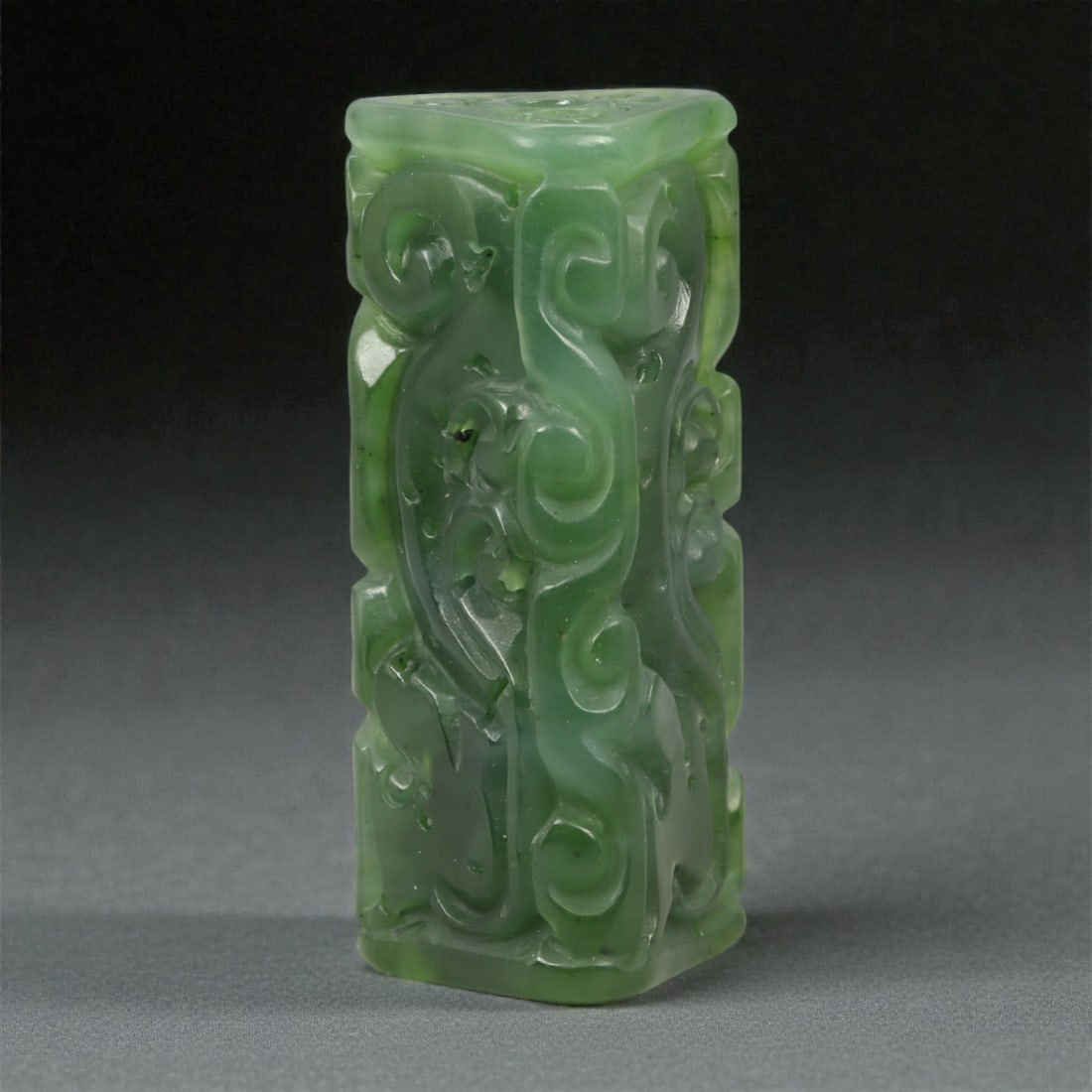 A Chinese Nephrite Jade Dragon-Form Bead, 20th Century, 7YA: Carved from translucent green nephrite jade with areas of darker saturation and minor natural inclusions, the object is formed as a rectangular bead worked in low relief on one face. The principal dec