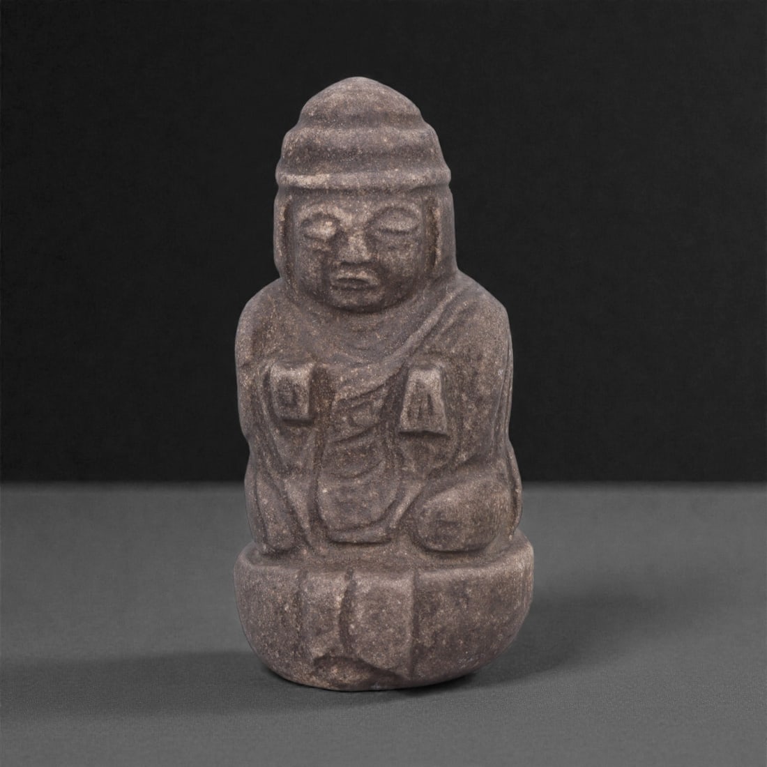 A Japanese Stone Buddhist Figure, Edo Period, 64W: Carved from dark grey stone with a fine granular texture and areas of surface wear, the figure represents a seated Buddhist figure rendered in compact, frontal form. The head is proportionally large,