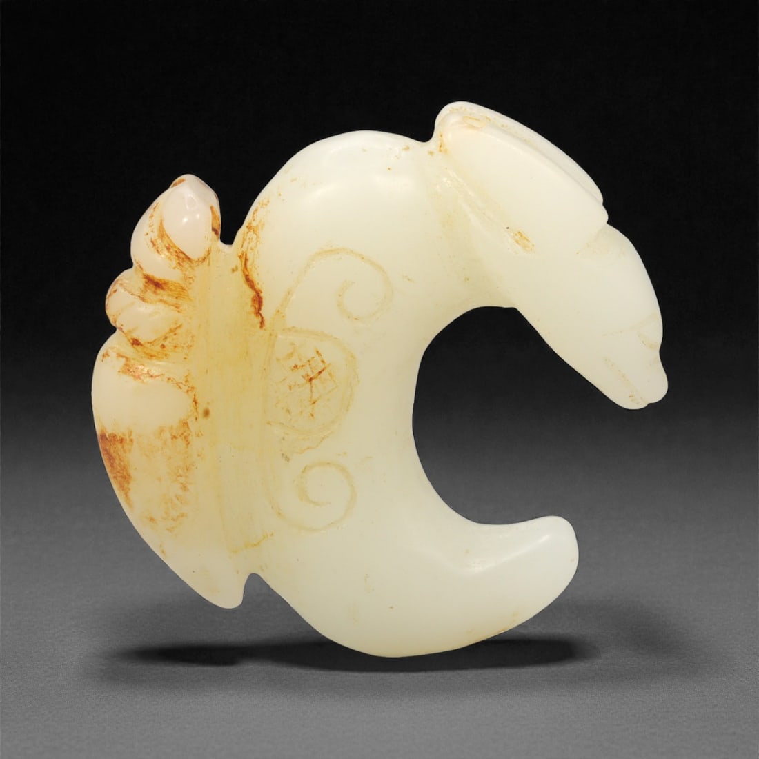A Chinese Hetian Jade Zoomorphic Ornament, 20th Century, Ancient Style, 83FH: Carved from pale creamy-white Hetian jade with areas of natural russet-brown staining, the ornament is worked in the form of a stylized animal rendered in a compact, curved silhouette. The head is cle