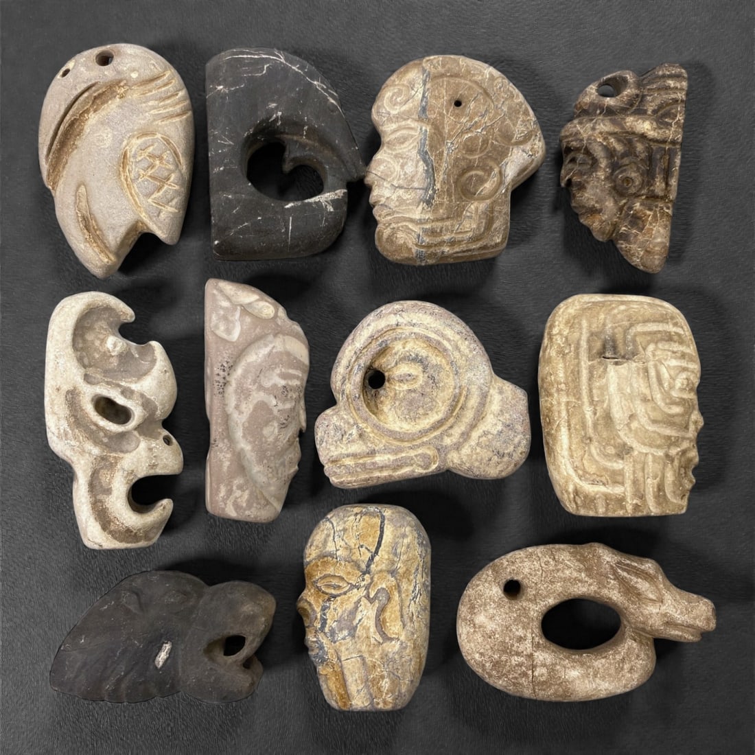 A Group of Mesoamerican Stone Sculptural Pendants, Pre-Columbian Period, 74Z: Carved from various stone types in grey, brown, and buff tones, the group comprises eleven sculptural pendants primarily representing stylized human heads and abstracted anthropomorphic or zoomorphic