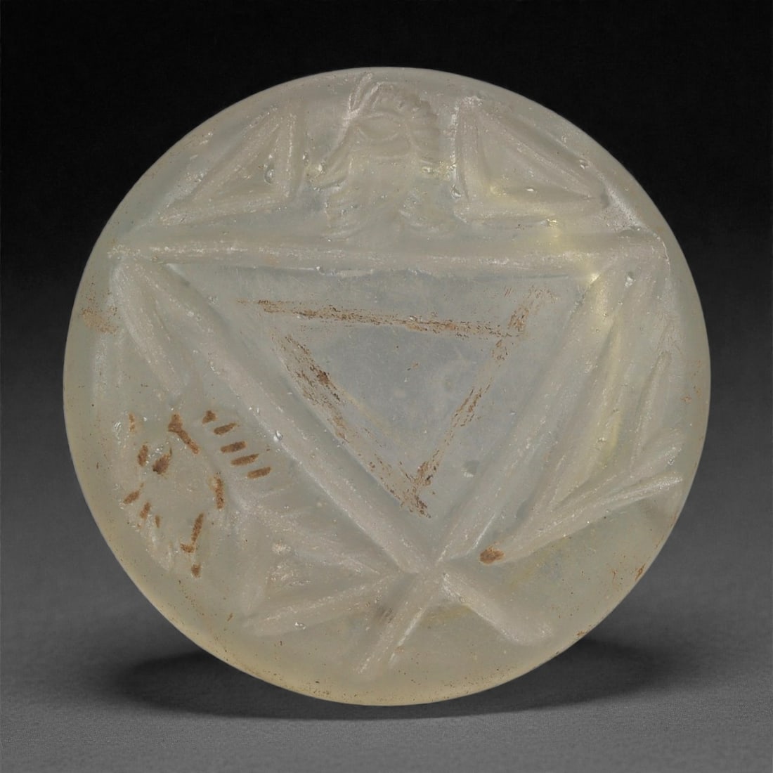 A Late Roman-Byzantine Glass Roundel with Anthropomorphic Motif, 3rd-6th C. CE, 26LP: Cast in pale translucent glass with a softly weathered surface, the roundel is molded in low relief with an anthropomorphic figure positioned above a large central triangle. The figure shows a schemat
