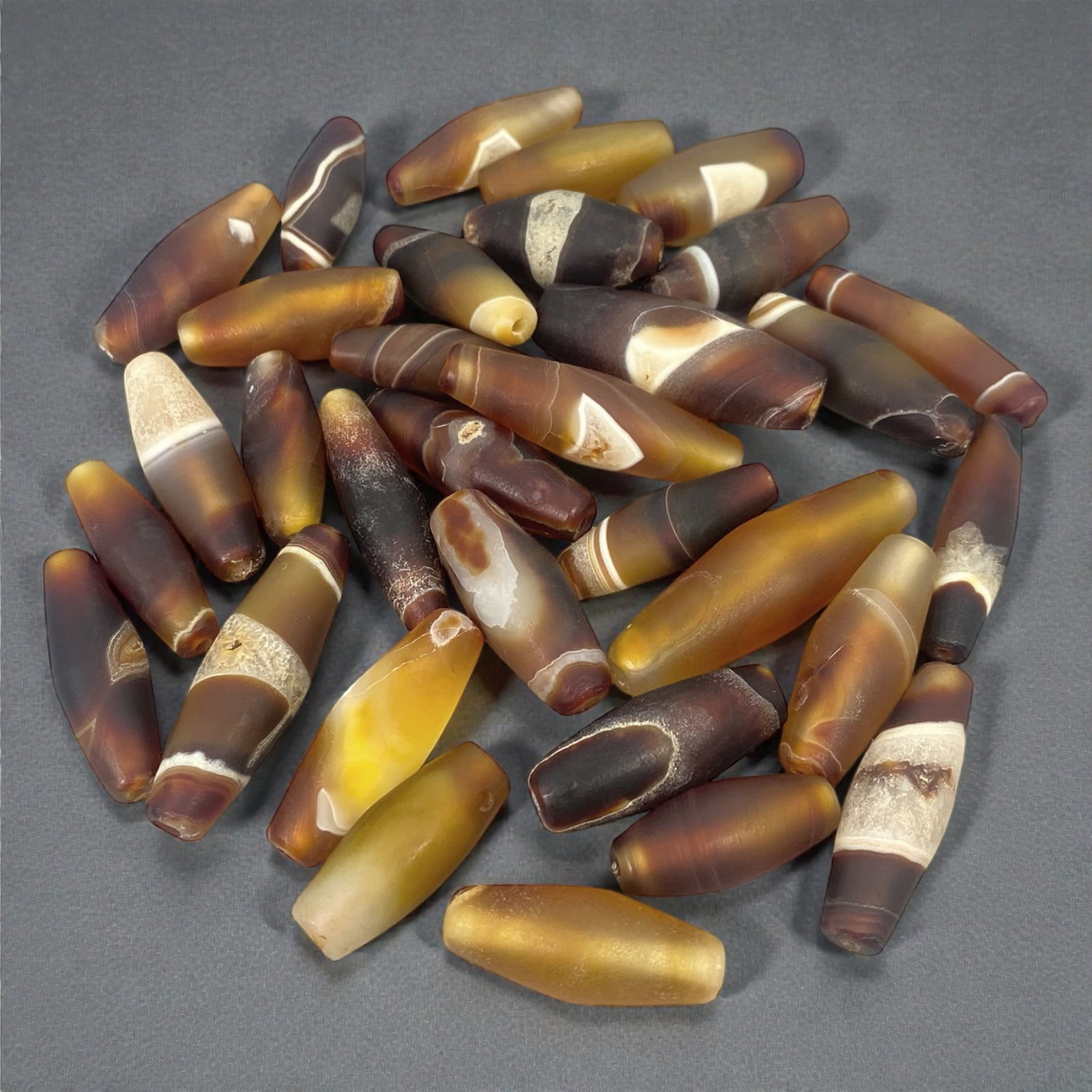 A Group of Himalayan Agate Chung Dzi Beads, 19th-20th Century CE, 11W: The group comprises thirty-two agate beads of elongated cylindrical and barrel forms, carved from translucent to semi-opaque chalcedony in tones of amber, honey, brown, cream, and dark grey. Several b