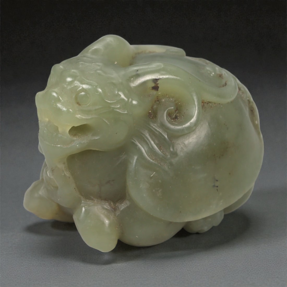 A Chinese Nephrite Jade Pixiu Figure, 20th Century, 3YA: Carved from pale green nephrite jade with soft translucency and scattered natural inclusions, the figure represents a pixiu shown in a compact, crouching posture. The head is strongly modeled with a p