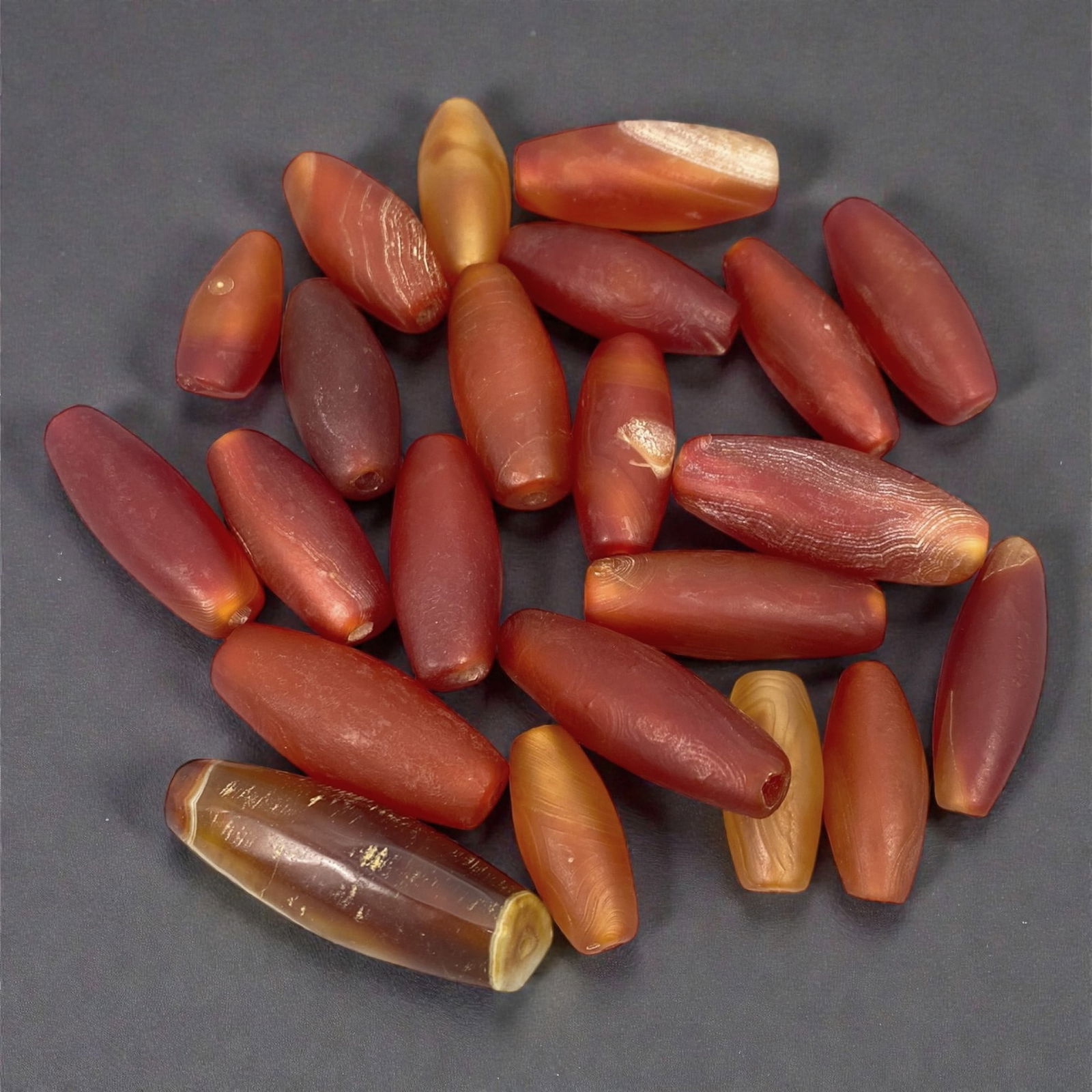 A Group of Tibetan Carnelian Beads, 18th-19th Century CE, 14W: The group comprises twenty-two carnelian beads of elongated barrel and tapering cylindrical form, carved from translucent to semi-opaque chalcedony in shades of deep red, orange-red, amber, and honey.