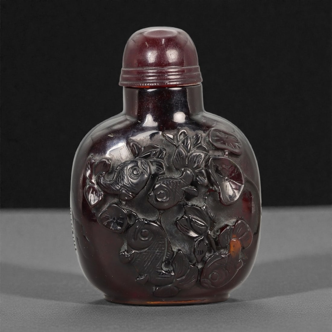 A Chinese Amber Snuff Bottle with Fish Motifs, 20th Century, 77FG: Carved from deep translucent amber of rich reddish-brown tone, the snuff bottle is of rounded rectangular form with softly bulging sides, rising from a narrow flat foot to a short cylindrical neck. Th