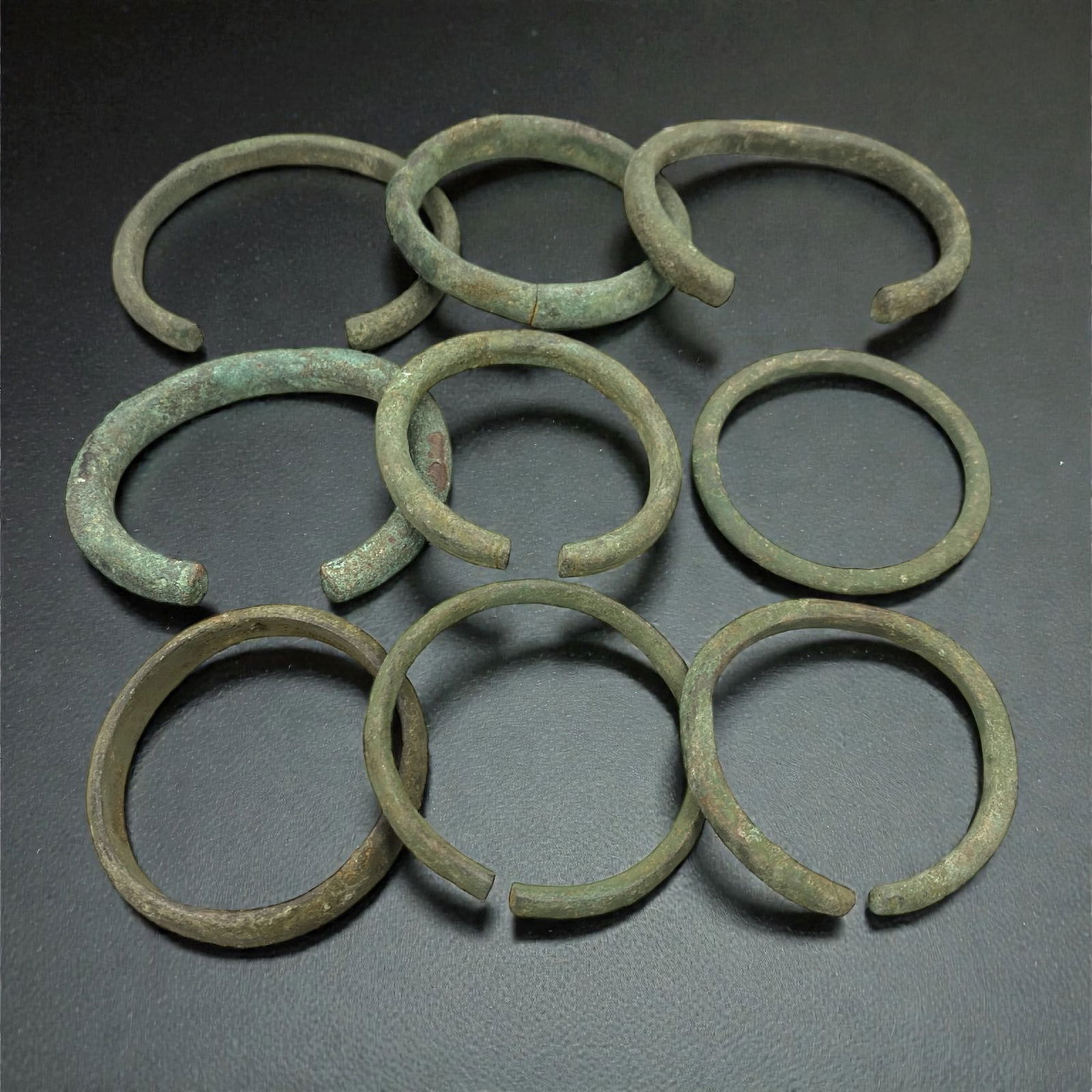 A Roman Bronze Bracelet Group, 1st-4th Century CE, 77W: The group comprises nine open-ended bracelets cast in bronze, each formed as a circular or near-circular hoop with rounded to slightly flattened cross-sections; the surfaces exhibit mottled green and