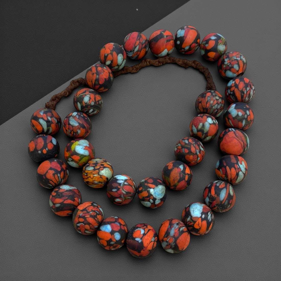 A West African Glass Mosaic Bead Necklace Strand, 19th-Early 20th Century, 265g, 9AA: Composed of spherical glass beads with a matte black ground decorated with irregular patches of vibrant red, orange, and pale blue, strung on a modern braided fiber cord. The mosaic pattern is charact