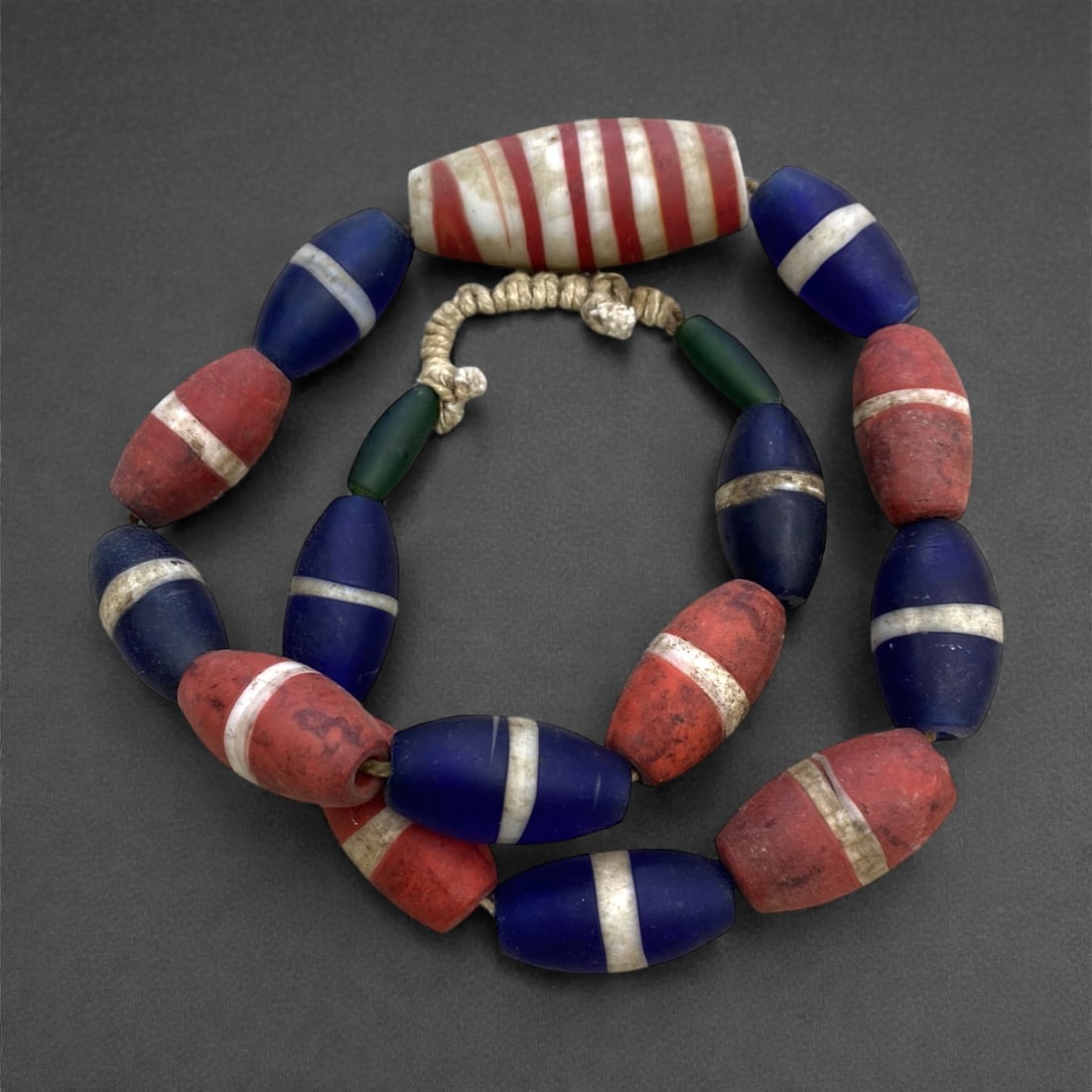 A Venetian Glass Trade Bead Necklace Strand, 19th Century, 281g, 23AA: Composed of elongated oval glass beads featuring bold single white stripes on red and blue grounds, crowned by a large central barrel bead with alternating red and white bands, strung on a period fibe