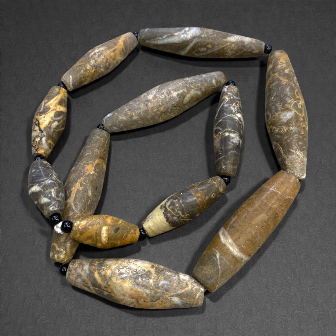 An Ancient Near Eastern Jasper Bead Necklace Strand, 122g, 28AA: Composed of elongated, hand-carved jasper beads in varying earthy tones of brown, grey, and ochre, interspersed with modern black glass spacers, strung on a contemporary cord. These beads exemplify th