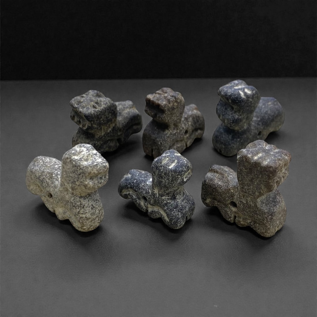 A Group of Six Bactrian Stone Feline Amulets, 3rd-2nd Millennium BCE, 37g, 53AA: Each amulet is carved from coarse-grained, dark stone, formed as a stylized recumbent feline with a compact body, raised head, and simplified facial features. A perforation runs horizontally through t